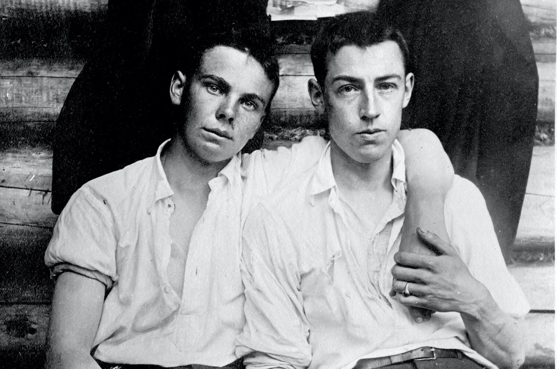 A black and white image of two men sitting in a cabin. The man on the left has his left arm around the shoulders of the man on the right. The man on the right is holding the other man's hand as it rests over his shoulder. He is wearing a ring on his left ring finger.